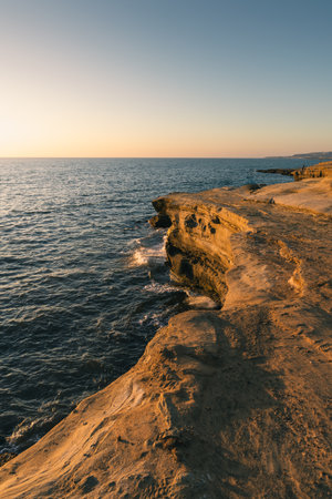 Waves gently crash against the rugged shoreline as the sun sets, casting warm colors over the landscape and water in the evening sky.の写真素材