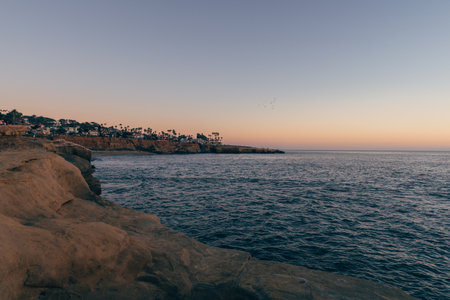 Soft colors fill the sky as the sun sets over calm waters. Cliffs and palm trees frame the serene coastal view, creating a peaceful atmosphere.の写真素材