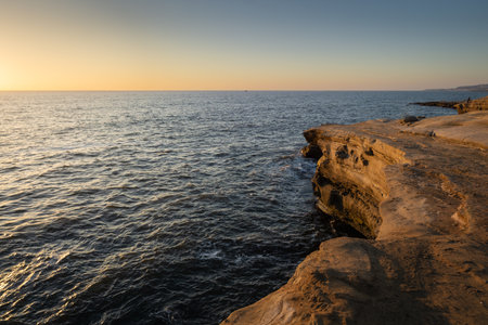 The sun sets over the calm ocean, casting warm colors across the sky while rocky cliffs line the...