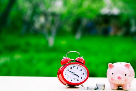 Pink piggy bank with red alarm cloack and coins on tableの写真素材