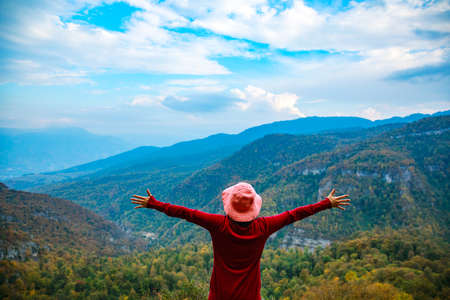 Back view of young hiker woman standing with raised arms under beautiful cloudy sky, enjoying fantastic view of mountains covered with pine forest.の写真素材