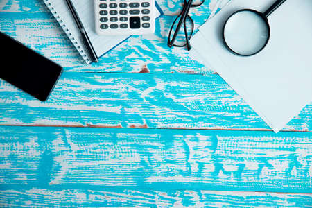 Magnifying glass on top of blank paper with multiple office supply on blue wooden table, search concept.の写真素材