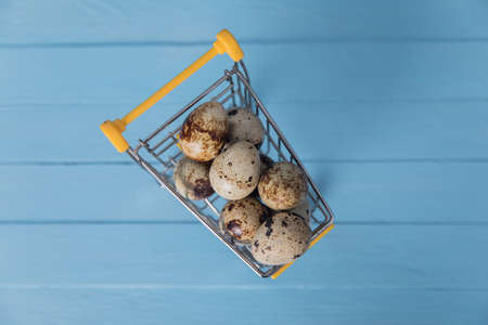 Raw quail eggs in mini shopping cart on blue bakground