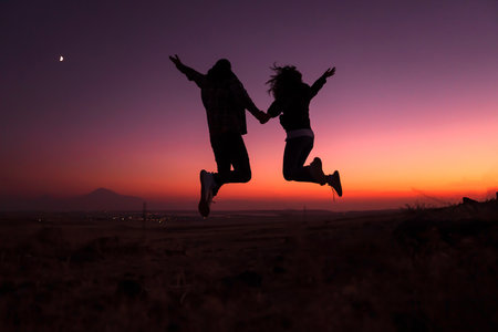 Silhouette of a couple jumping against the sky during sunset on a hillの写真素材