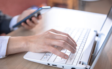 Woman typing on laptop while holding phone in officeの写真素材