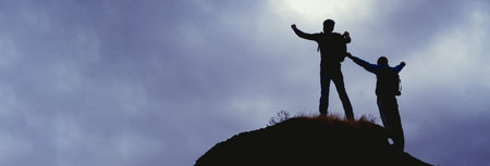 Silhouettes of two mountain climbers on the top of a mountain with victorious gesture.の写真素材