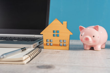 Laptop, Wooden Cabin, and Piggy Bank on the Office Table, home sale conceptの写真素材
