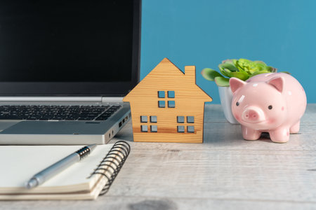 Laptop, Wooden Cabin, and Piggy Bank on the Office Table, home sale conceptの写真素材