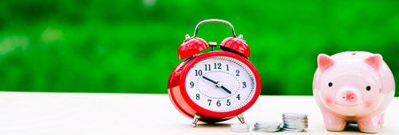 Pink piggy bank with red alarm cloack and coins on tableの写真素材