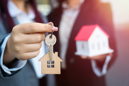 Two business women, one holding a small wooden house, the other a key, house sale, business conceptの写真素材