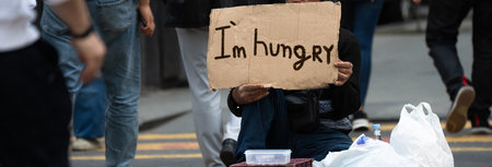 A beggar on a city street holding a cardboard with I'm hungry written on it