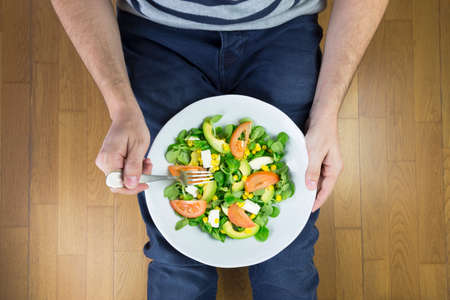 Young man in jeans eating tasty salad with vegetables while sitting on the floor. Healthy food...