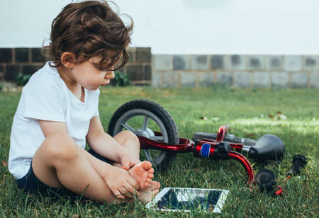 A little boy sitting on the green grass and looking at a digital tablet with a red bike.