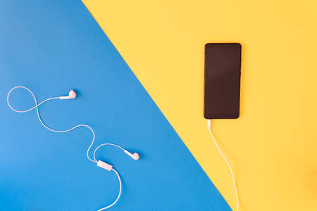 Smartphone and earphones on blue and yellow background top view. Headphones and device on colored background. Smartphone with earphones on color backgroundの写真素材