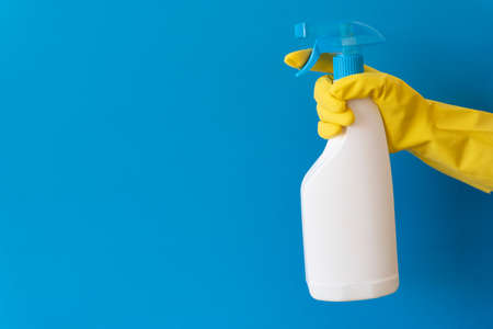 Hand in a yellow rubber glove holds cleaner spray bottle over blue background. Cleaning service...