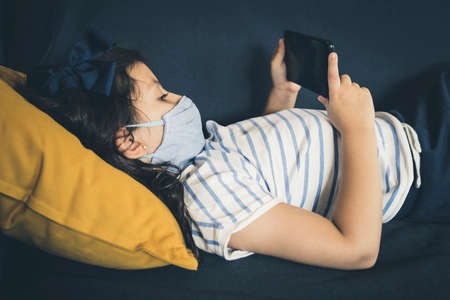 Close up of a relaxed young girl with mask using a Smartphone lying on a sofa in the living room at...