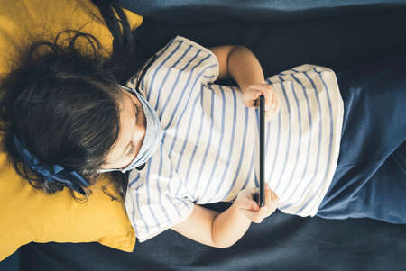 Close up of a relaxed young girl with mask using a Smartphone lying on a sofa in the living room at home with a cozy background during the Coronavirus pandemic. Top view.の写真素材