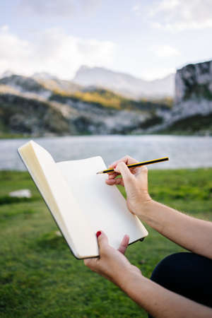 A beautiful caucasian blonde woman writes and draws on a notepad in a mountain landscape with lakeの写真素材
