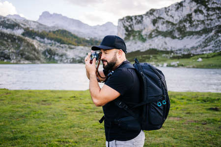 Caucasian man with cap and beard makes photos with a photo camera sightseeing in a mountain landscape with a lakeの写真素材