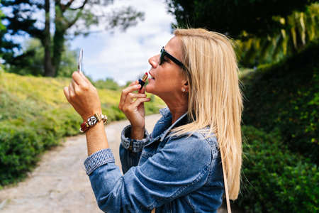A beautiful caucasian blonde woman puts on makeup on the street in a park