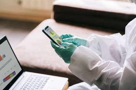The hands of a person using the mobile phone with gloves and a safety suit against a pandemic or virusの写真素材