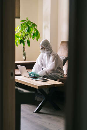 A woman works from home with the laptop, digital tablet and mobile phone in the living room, equipped with security measures against a virus prevention with a safety suit, glasses, mask and latex gloves.の写真素材