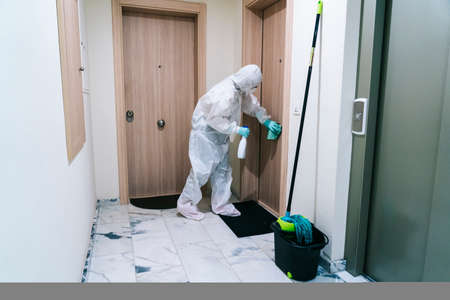 A person wearing a mask, gloves and a safety suit cleans and disinfects a doorway of a community flat in the face of a virus pandemic, protected safety equipment so as not to be infectedの写真素材