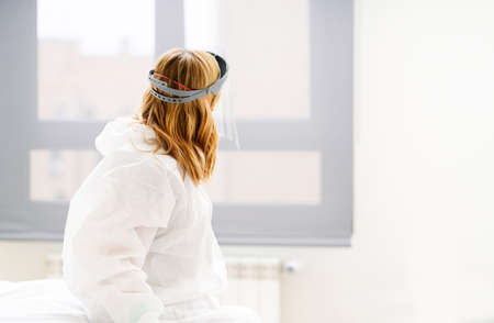 A female doctor on her back sitting on a hospital bed looking towards the window wearing a safety suit and protective maskの写真素材