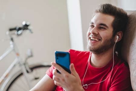 A boy in a red T-shirt uses the mobile phone with headphones relaxed and happy on the sofa at home. Stay at home.の写真素材