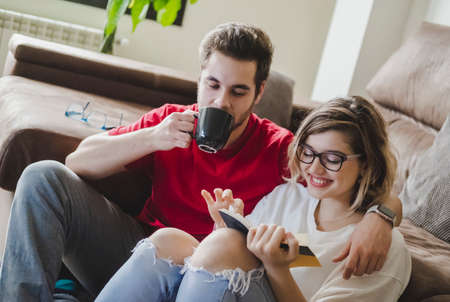 A couple together sitting on the floor in the living room at home, drinking coffee and reading a...