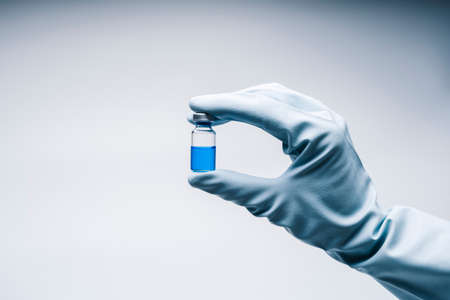 A hand with a latex glove holds a medical vial for injection with syringe with liquid of medicine or...