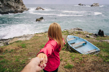 A blonde woman leads her partner by the hand to a cove with sea and rocks with an abandoned wooden boat. Rear view of woman wearing pink coat.の写真素材