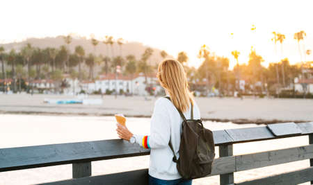 A blonde woman in glasses enjoys ice cream at sunset on a California pier with palm trees in the...