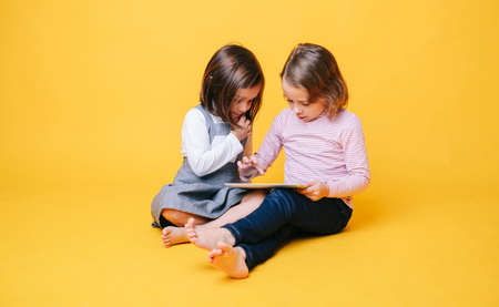 Two girls use a digital tablet on an isolated yellow background with copy spaceの写真素材