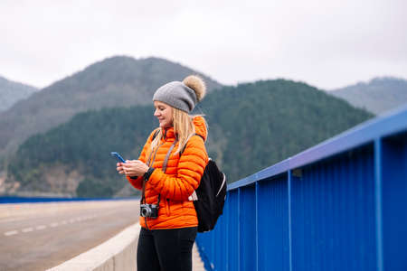 Side view of a woman in orange coat and wool hat using a mobile phone on a road with mountains in...