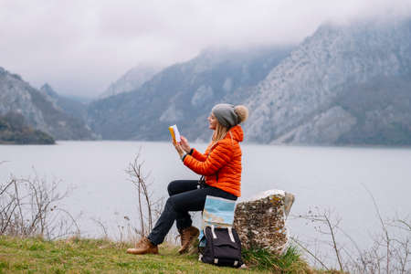 Side view of woman in a wool hat and orange coat reads a book in nature with map and bagの写真素材