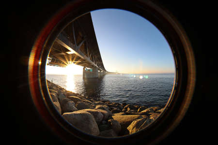 Sun seen through the Oresund bridge at the viewpoint near Limhamn, Sweden, in February 2021. Wide angle, clear sky, bridge stretching from the left to the horizon. Lens flare, sun approaching sunset and seen under the bridge between its pillars.の写真素材