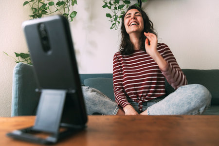 Girl laughing looking at the phone sitting on the sofa at home. Selective focus on the girl. Stay at home conceptの写真素材