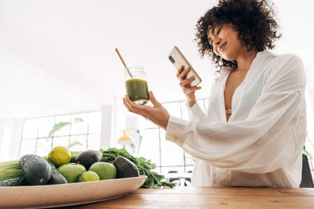 Young african american woman taking photo of green juice and vegetables with cellphone at home. Home concept. Technology concept. Health concept. Copy spaceの写真素材