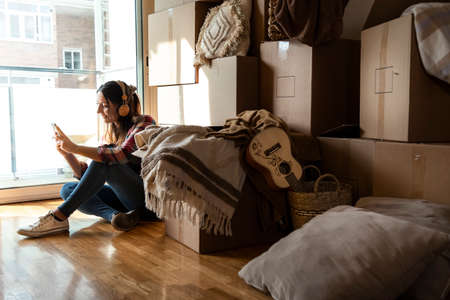 Moving in. Woman listening to music with headphones looking at cellphone next to pile of cardboard...
