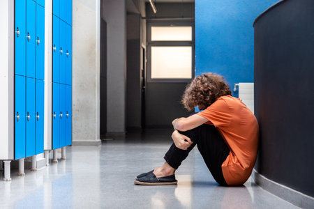 Sad caucasian teen boy victim of bullying sitting on floor in high school corridor. Copy space. Bullying consequences concept. Sadness and depressionの写真素材