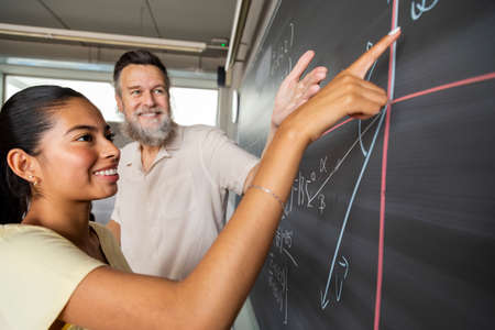 Happy high school teen girl student shows teacher maths problem solution pointing with finger to...