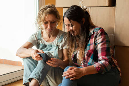 Two women look at cellphone next to a pile of moving cardboard boxes. Relocation concept. Moving in.の写真素材