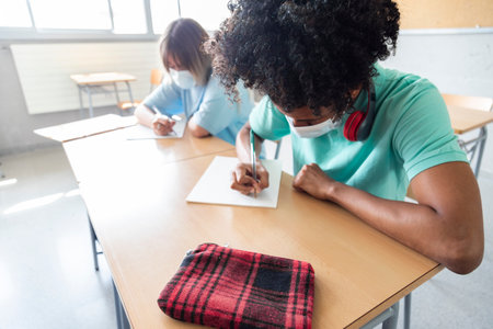 Black teen boy wearing face mask in class. High school student writing on paper. Copy space. Educaction concept. Healthcare concept.の写真素材