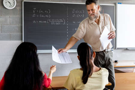 Mature adult caucasian male teacher handing out sheet of paper to two teenager girl high school students. Education concept.の写真素材