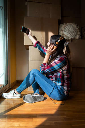 Young woman taking selfie with headphones next to pile of cardboard boxes. Moving out concept....
