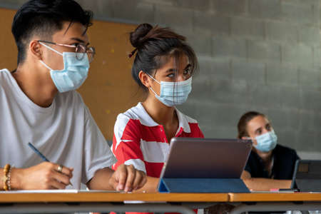 Close up of teen asian girl student in class wearing a face mask using tablet. Multiracial classmates. Education concept. Healthcare concept.の写真素材