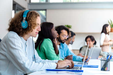 Young caucasian male with headphones works in spacious coworking office. People working in background. Copy space. Coworking concept. Startup concept.の写真素材