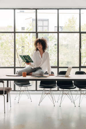 Young mixed race woman reading journal. Sitting on desk. Carefree and happy student. Messy desktop....