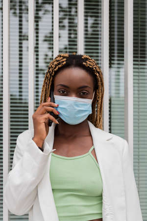 Portrait of young african american woman looking at camera talking with cellphone wearing a face mask. Black woman with blond braids using her mobile phone. Vertical image. Technology concept. Health care concept. Copy spaceの写真素材